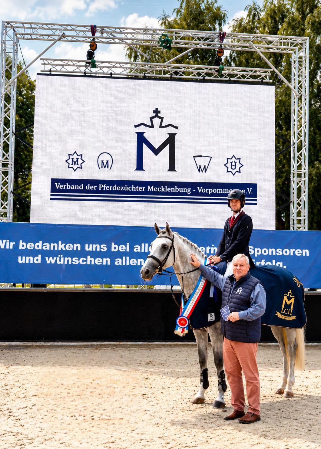 weißer Hengst mit Siegerdecke und Scherpe davor ein Gratulant